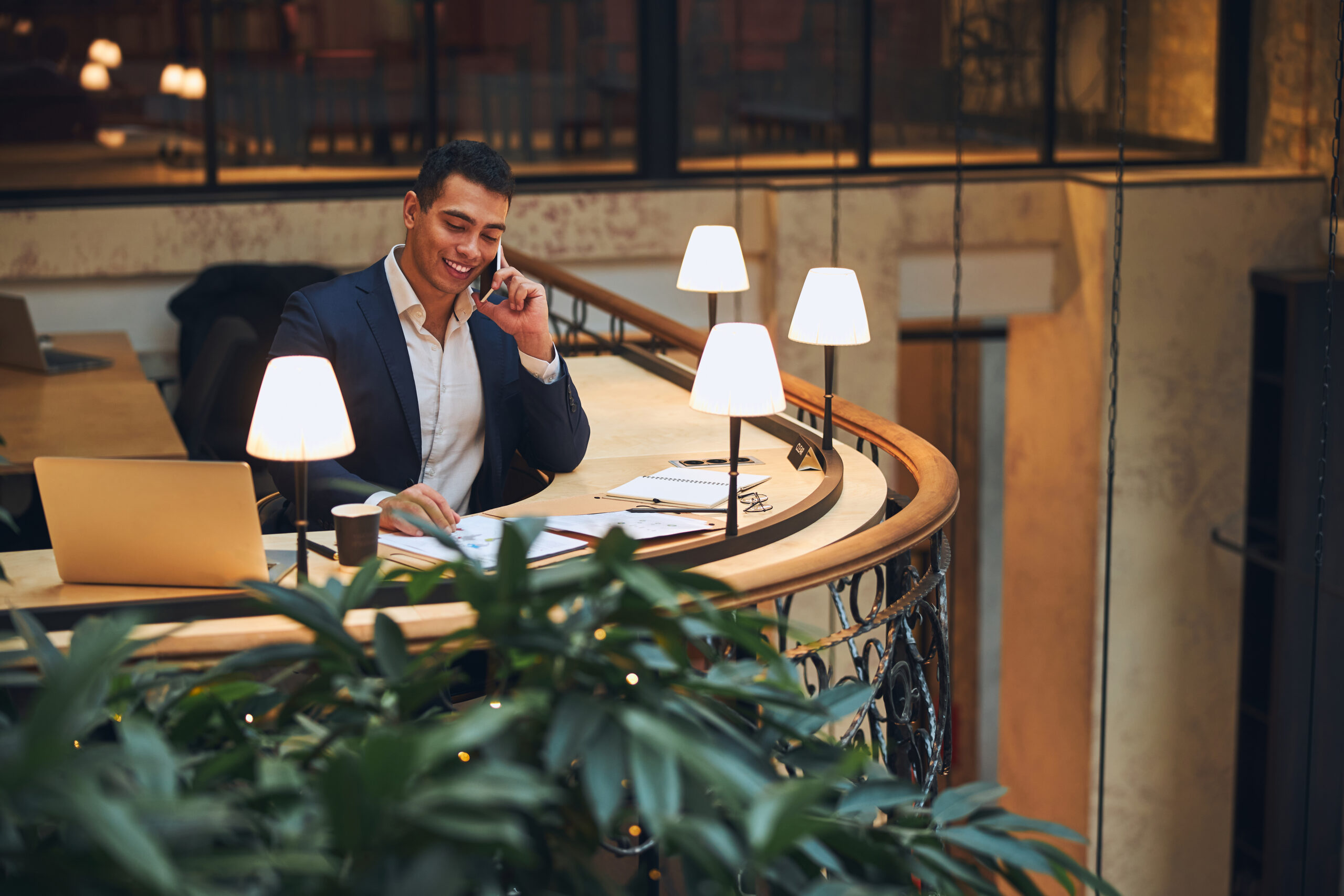 Waist-up portrait of a smiling businessman looking at the documents on the desk during the phone conversation